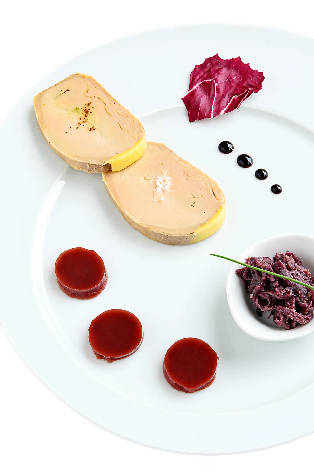 Deux tranches de foie gras dressées sur une assiette blanche avec trois palets de gelée rouge, un petit bol de chou rouge, une feuille de radicchio et des gouttes de réduction balsamique.