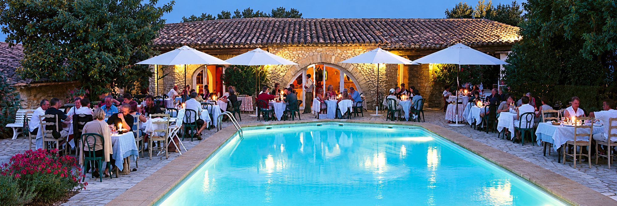 Des convives dînent à la tombée de la nuit sur une terrasse éclairée autour d’une piscine, devant un bâtiment en pierre et sous des parasols blancs.