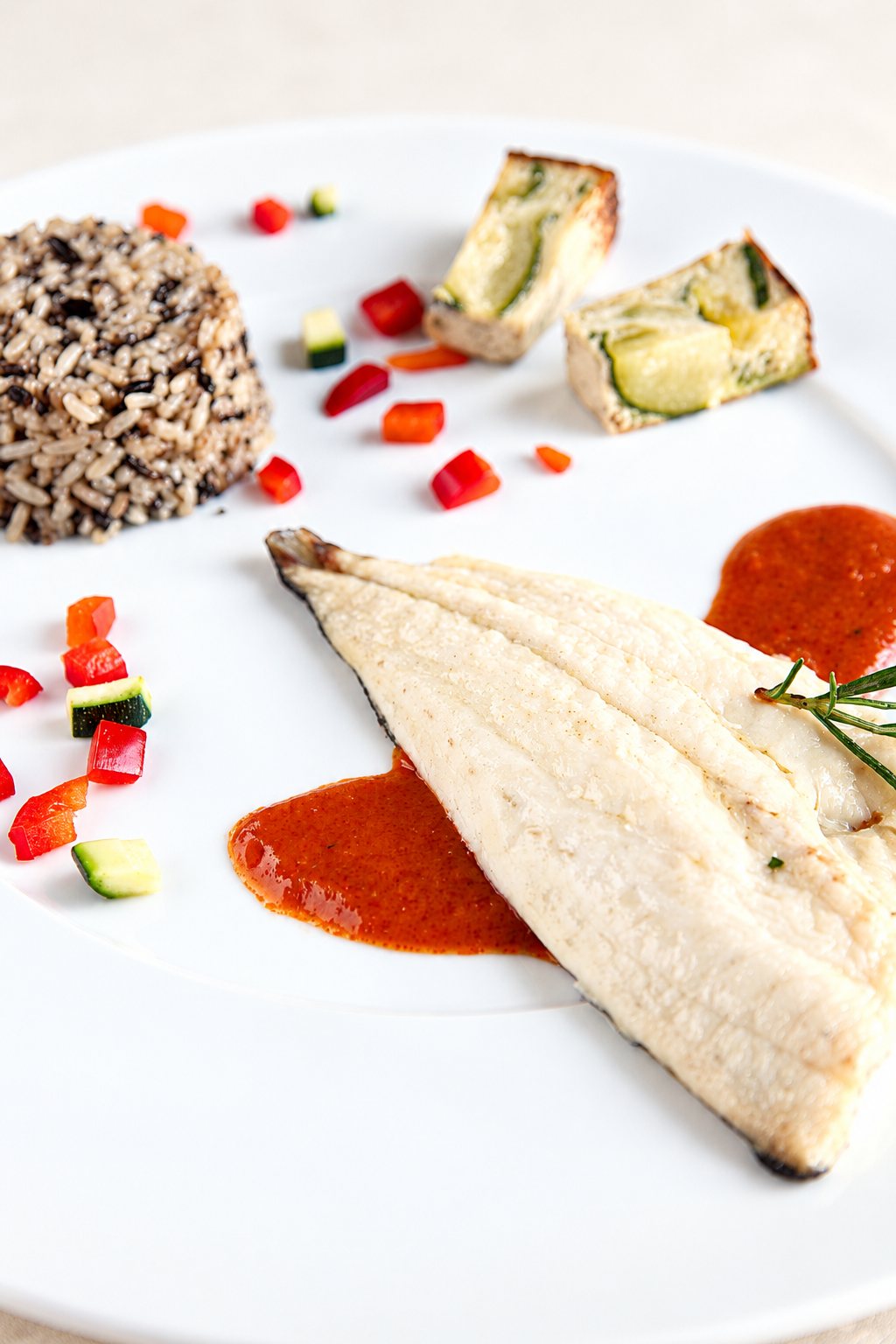 Assiette blanche garnie d'un filet de poisson avec une sauce rouge, du riz sauvage, des dés de poivron et de courgette et un brin de romarin.