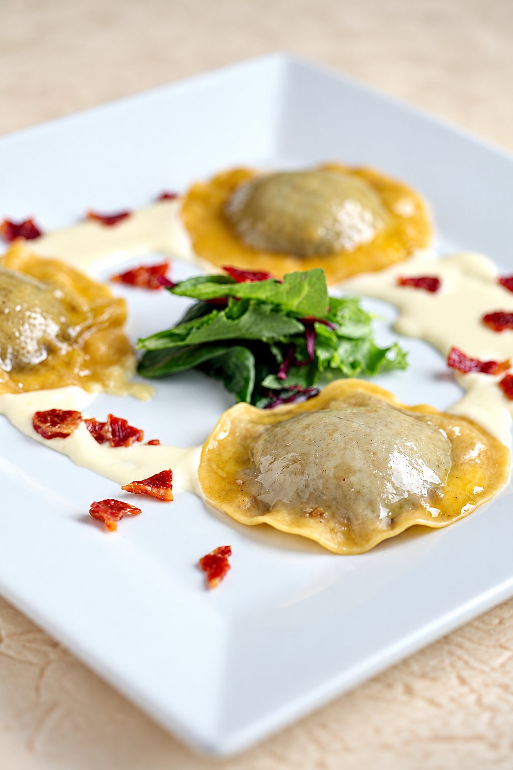 Trois raviolis dorés dans une assiette carrée blanche, accompagnés de crème, de jeunes pousses de salade et d'éclats de jambon séché croustillant.