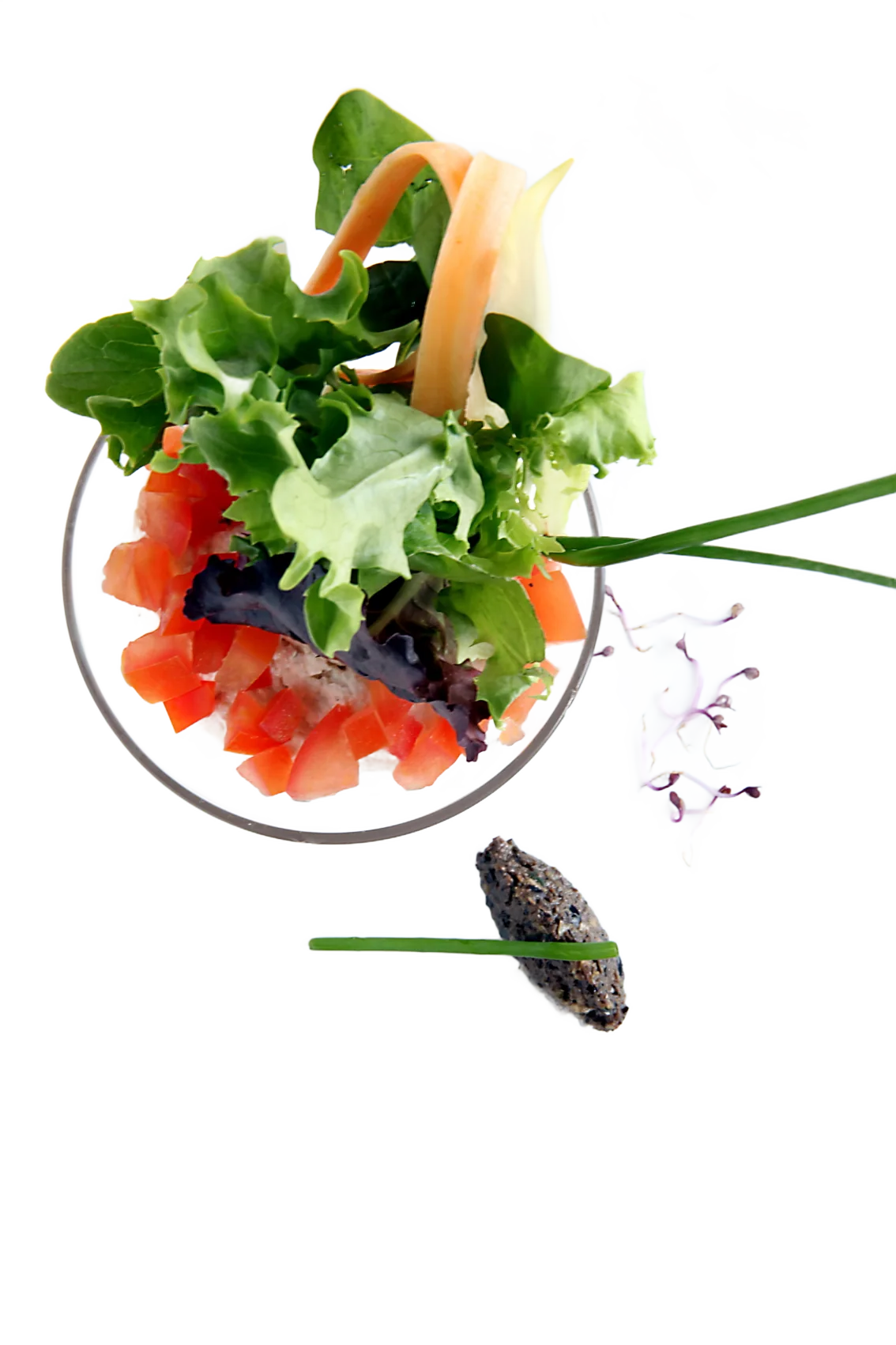 Vue du dessus d'une verrine de salade verte, dés de tomate et lanières de carotte, posée sur une assiette carrée blanche avec une quenelle de tapenade et des pousses roses.